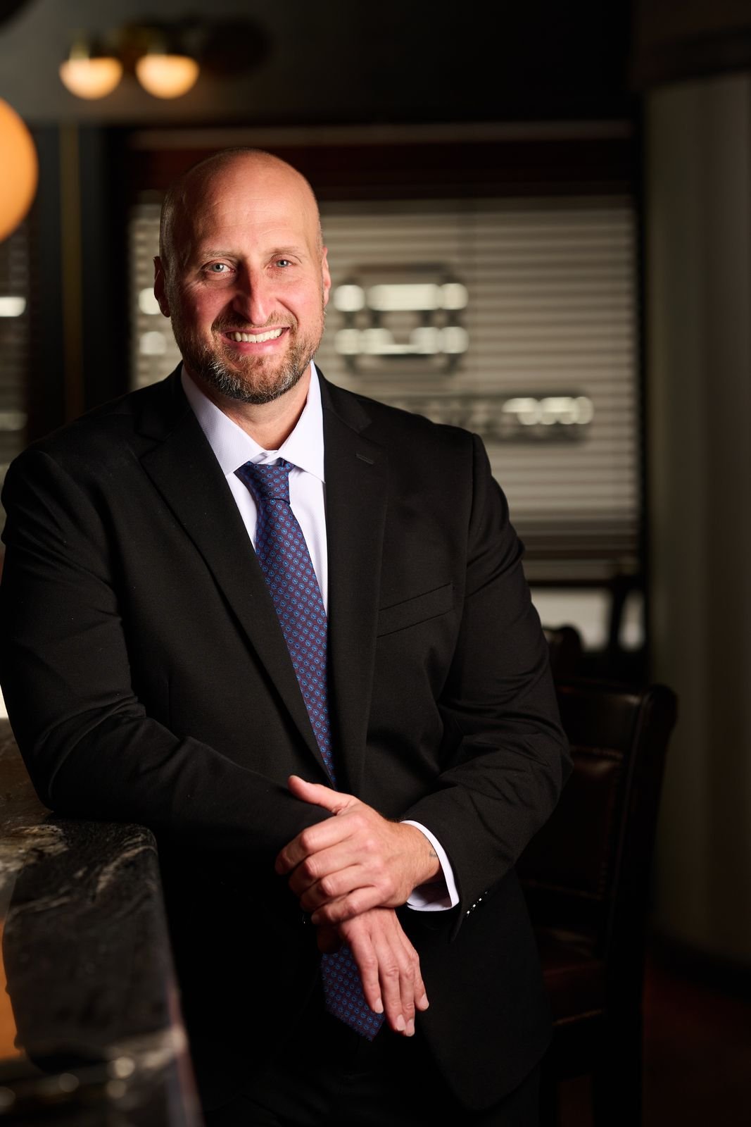 Dennis Turcinovic in a dark suit and patterned tie, smiling, leaning on the marble bar at Delmonico's