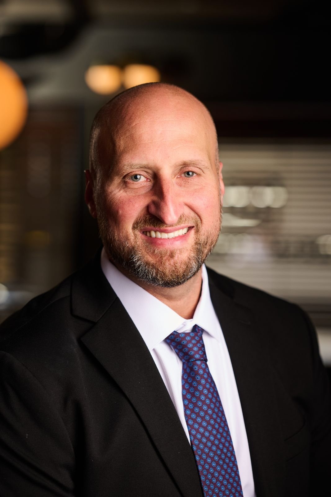 Portrait of Dennis Turcinovic in a navy suit and blue tie, smiling, in the warm light of the Delmonico's bar