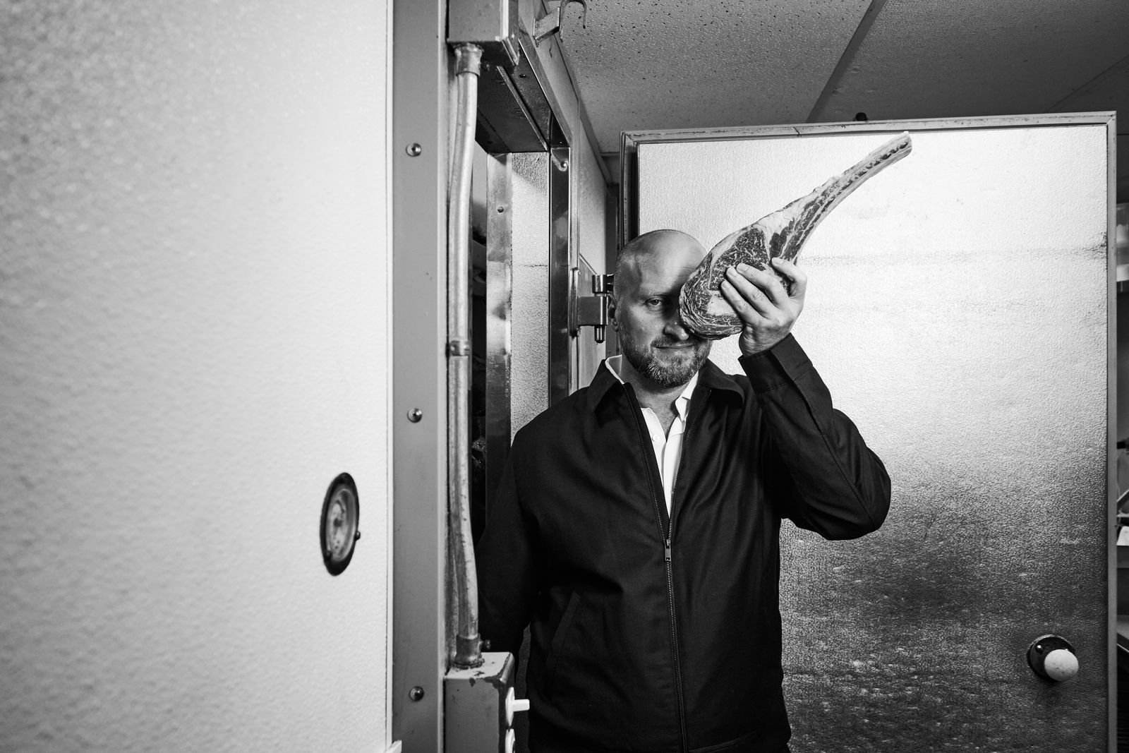 Black-and-white photo of Dennis Turcinovic at the walk-in door of the Delmonico's kitchen, holding a tomahawk steak